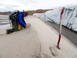 Research Kurt Kjær kneels on all fours on a ridge of sand in front of the glacier's edge.